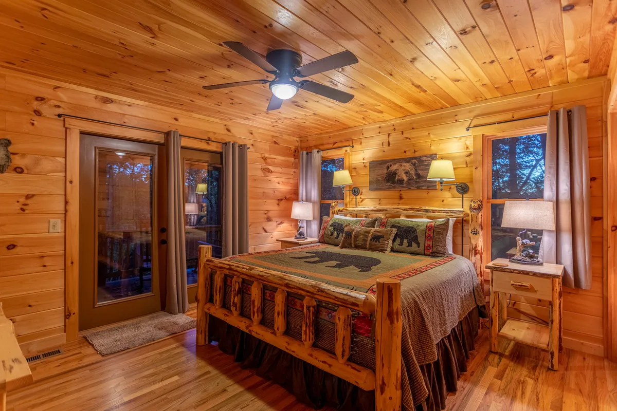 This image showcases a king-sized log bed adorned with bear-themed bedding, a nightstand with a lamp, and rustic wood paneling. Natural light filters in from a curtained window, creating a cozy, private retreat.