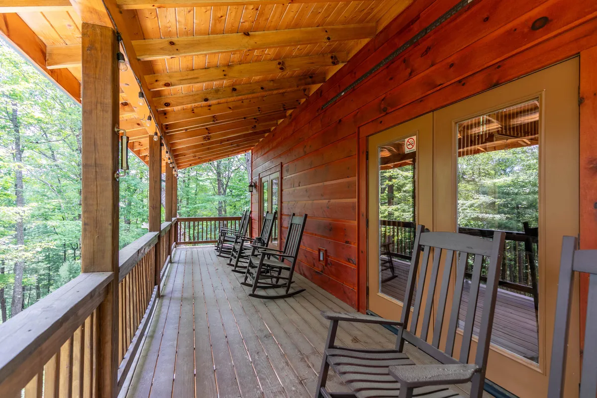 A covered deck with multiple classic rocking chairs, wooden railing, and lush greenery all around. The scene invites leisurely mornings or peaceful sunset moments overlooking the property.