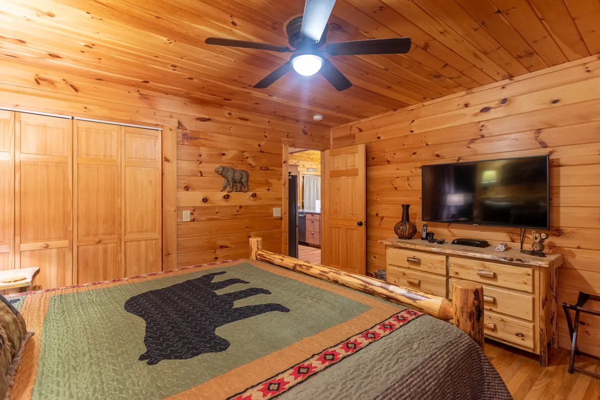 A warm and inviting bedroom with a log bed accented by a bear-themed quilt, a wooden dresser, and a flat-screen TV. The soft lighting and earthy decor create a comfortable, private retreat.