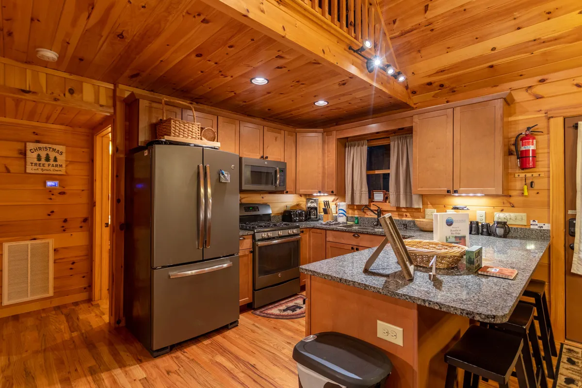 This image features a welcoming kitchen with granite countertops, stainless steel appliances, wooden cabinets, and a window with curtains bringing in natural light. Bar stools line the breakfast bar, creating an inviting spot for casual meals or conversation while cooking.
