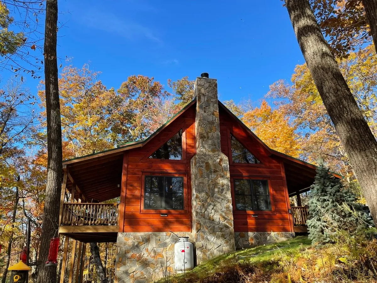 A charming wooden cabin surrounded by trees, featuring a stone chimney and a spacious deck. The setting is serene and private, offering ample outdoor space for relaxing, family gatherings, and nature walks. The deck provides a direct view of the natural landscape, making it ideal for enjoying the outdoors with pets.