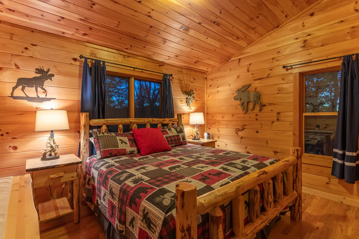 A charming bedroom with a log bed dressed in plaid bedding, moose decorations, and a nightstand with a lamp. The space feels both inviting and distinctly rustic, perfect for quiet moments or reading before bed.