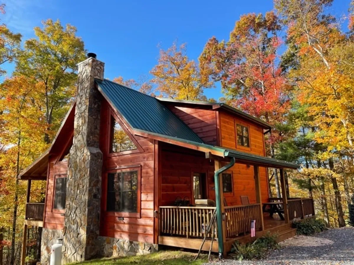 A broad perspective of the wooden cabin exterior, with a prominent stone chimney, colorful autumn foliage, and visible outdoor amenities. Groups are shown relaxing on the porch and gathered near a fire pit, embodying the spirit of casual outdoor living.
