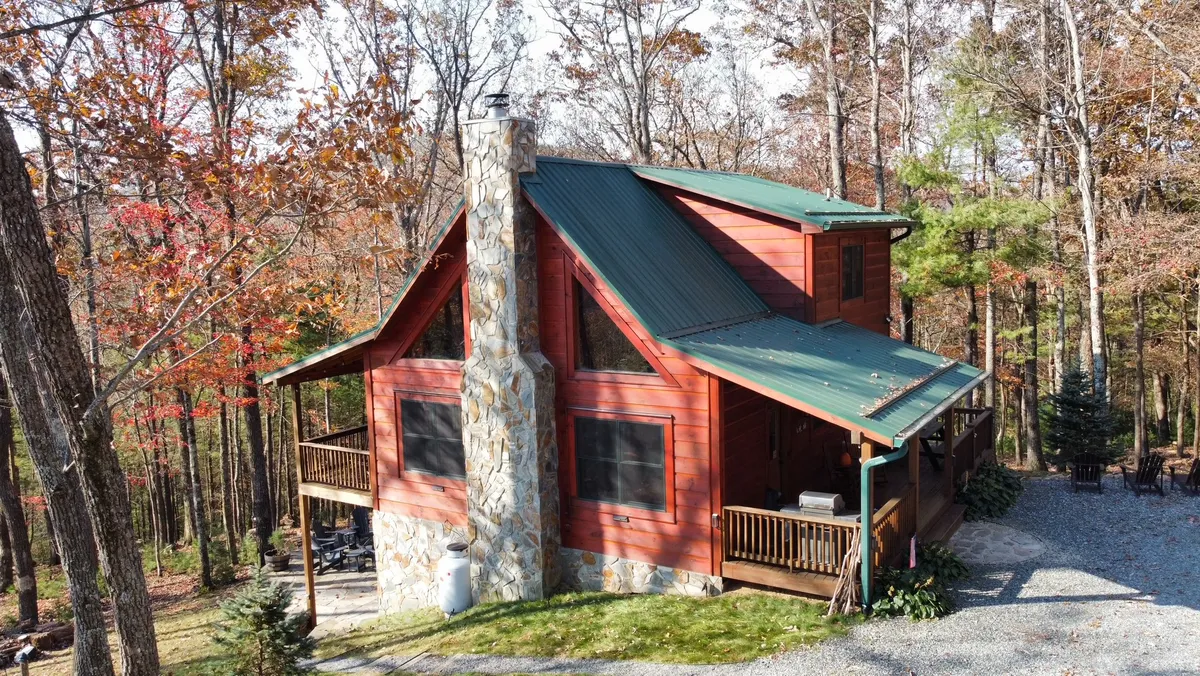 A charming log cabin with a classic stone chimney and inviting wooden deck, nestled among towering trees on over four private acres. The scene radiates tranquility, with sunlight filtering onto the deck—perfect for relaxing or gathering at sunset—and hints of a cozy fire pit area ideal for evening conversations under the stars.