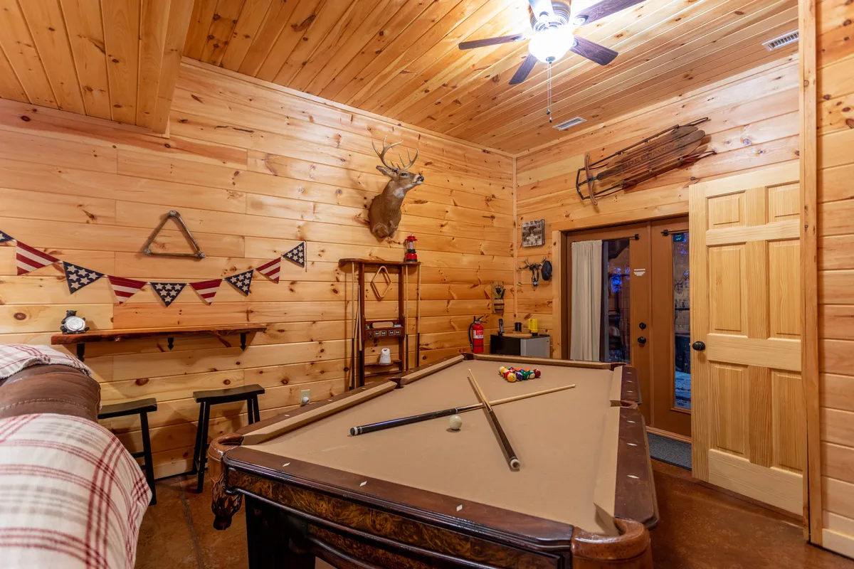 A rustic game room with wood-paneled walls centers on a pool table, with a mounted deer head and festive bunting adding to the friendly, cabin-like atmosphere. The space is ready for friendly competitions and post-adventure relaxation, ideal for groups unwinding together after a day outdoors.