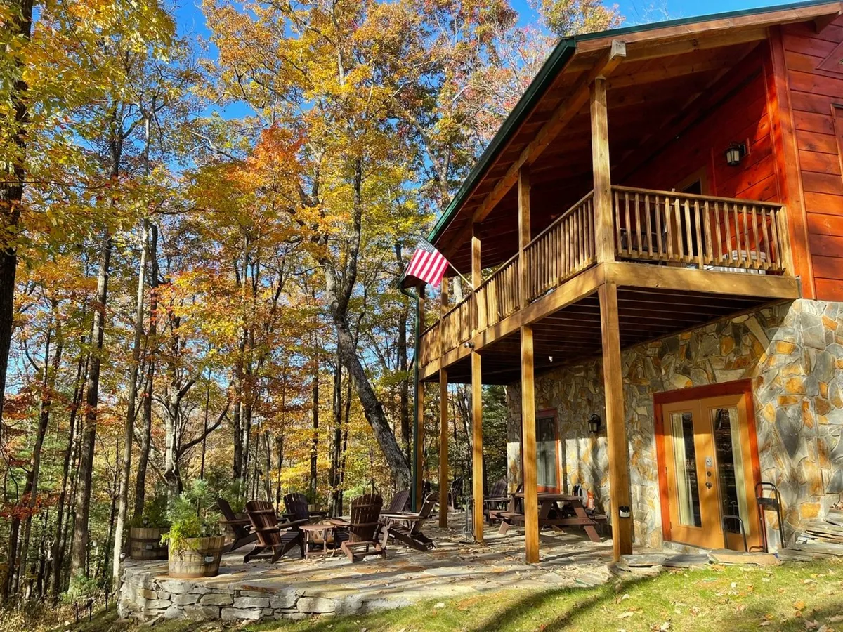 A spacious wooden deck surrounded by towering trees, featuring Adirondack chairs and an American flag. The deck offers a scenic vantage point for outdoor gatherings, barbecues, and quiet moments in nature.