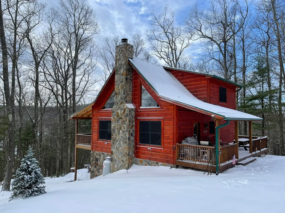 A classic log cabin with a stone chimney sits nestled among trees, its spacious deck visible and inviting. There is a light dusting of snow on the ground and the cabin itself, hinting at year-round adventure and the area's winter charm. The deck is large enough for gatherings and offers a connection to the natural surroundings.