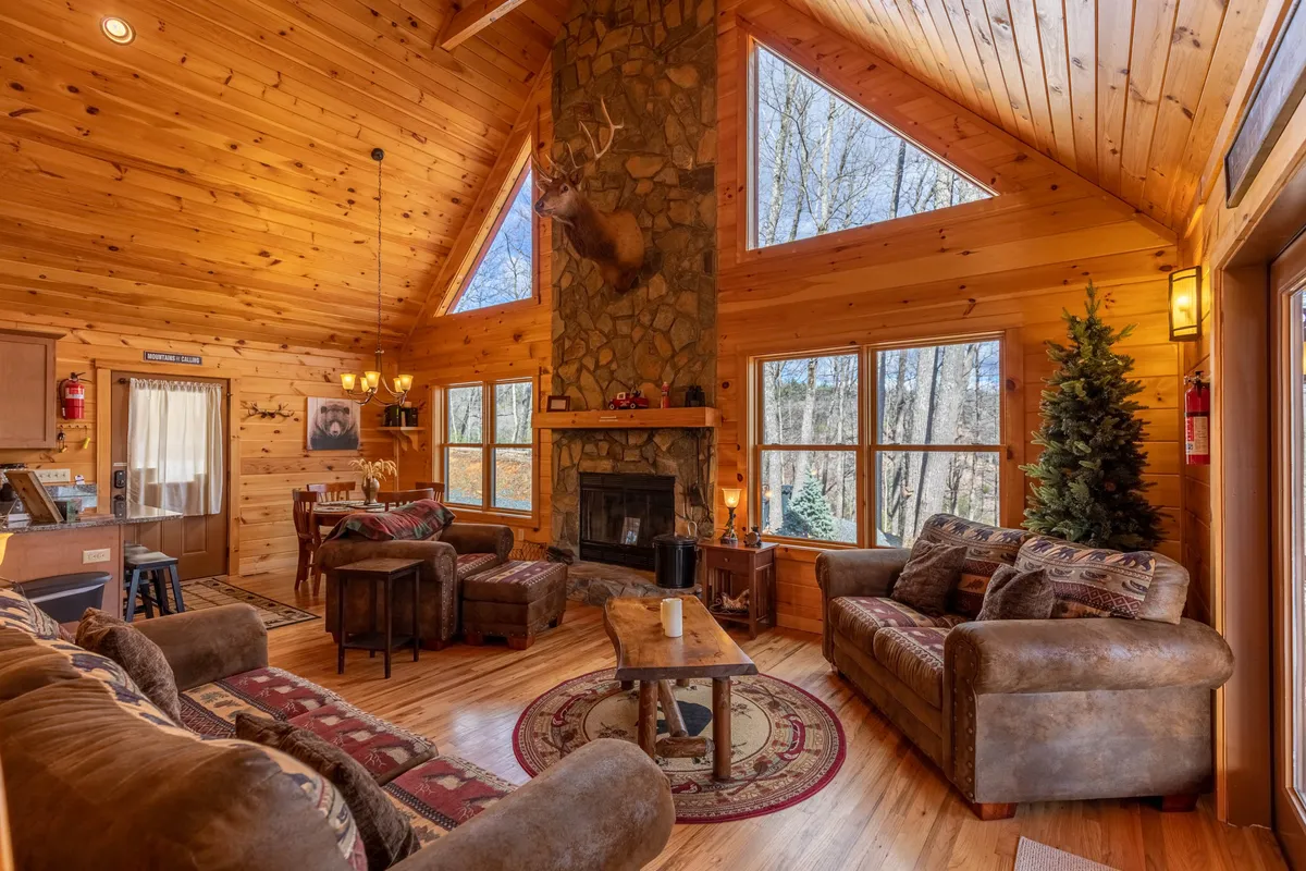 A spacious open-concept living area featuring a grand stone fireplace as its centerpiece, surrounded by plush sofas and a rustic wooden dining table. Large windows invite natural light and offer scenic views, while the cozy arrangement encourages gatherings and relaxed conversation.